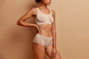 Sideways shot of slim woman wears cropped top and panties demonstrates perfect figure after weigh loss poses against beige studio background