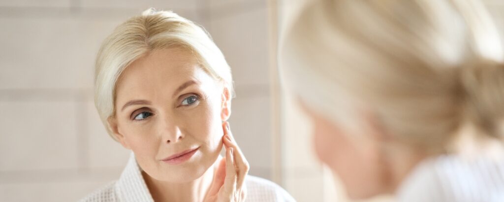 Headshot of gorgeous mid age adult 50 years old blonde woman standing in bathroom after shower touching face, looking at reflection in mirror doing morning beauty routine. Older skin care concept.